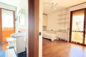 Een badkamer met toilet, wastafel en spiegel, en een slaapkamer met bed, planken en een raam.