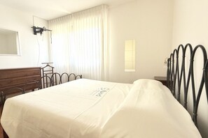 Hotelkamer met een bed, een dressoir, een spiegel, een raam met gordijnen en een metalen hoofdeinde.