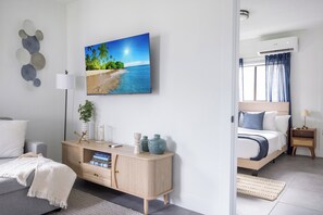 Un dormitorio moderno con cama, mesita de noche, lámpara y televisor de pared mostrando una escena de playa.