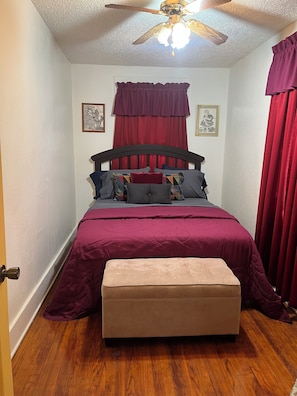 A bedroom with a bed, a ceiling fan, a window with red curtains, and a wooden floor.