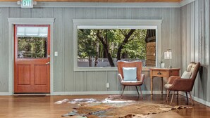 Une pièce avec une porte en bois, une grande fenêtre donnant sur des arbres, un bureau, deux chaises et un tapis.