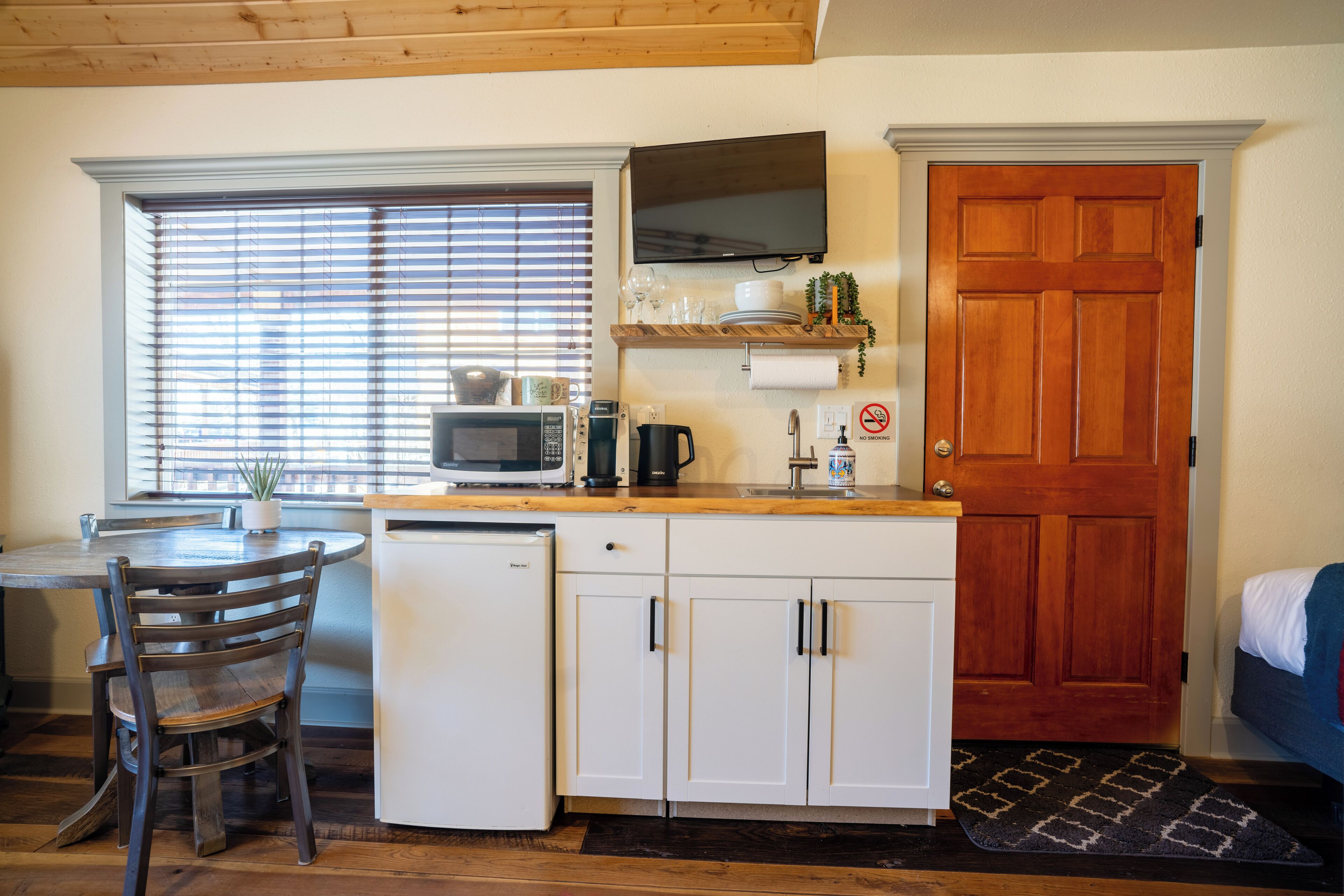 A compact kitchen with a microwave, coffee maker, and a sink. There's a dining area with a table and chairs, and a wooden door.