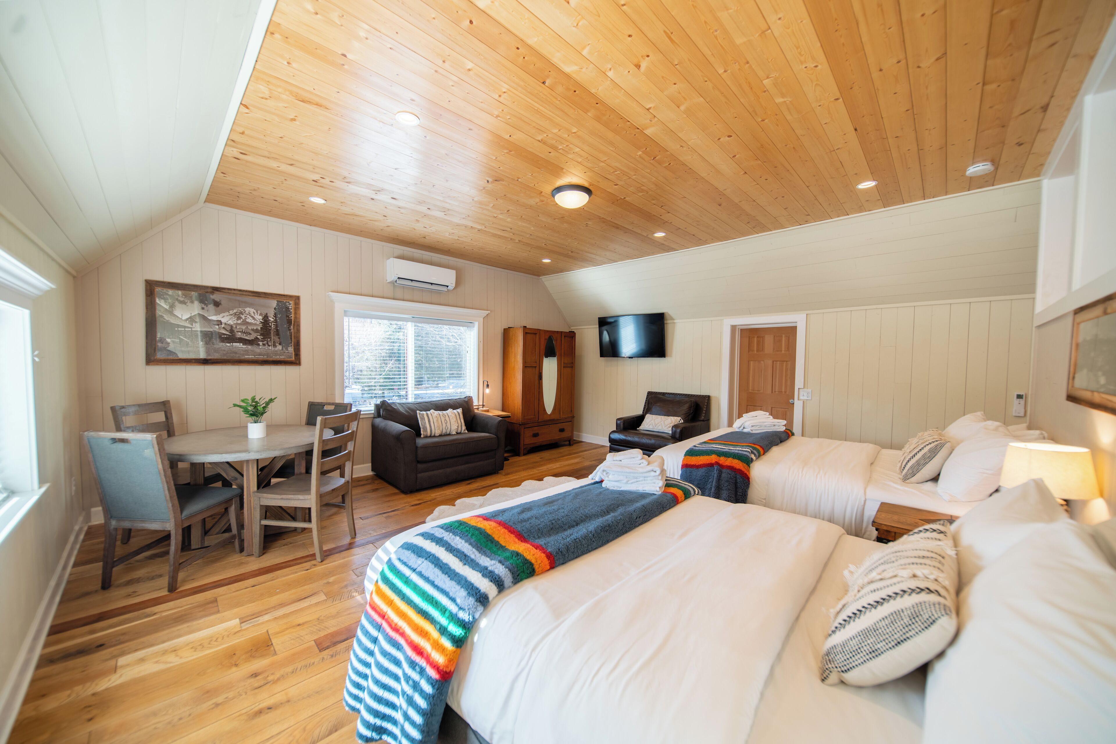 Ruime kamer met houten plafond, eethoek met tafel en stoelen, een bank, een aan de muur bevestigde televisie en een bed met strepenbedekking.