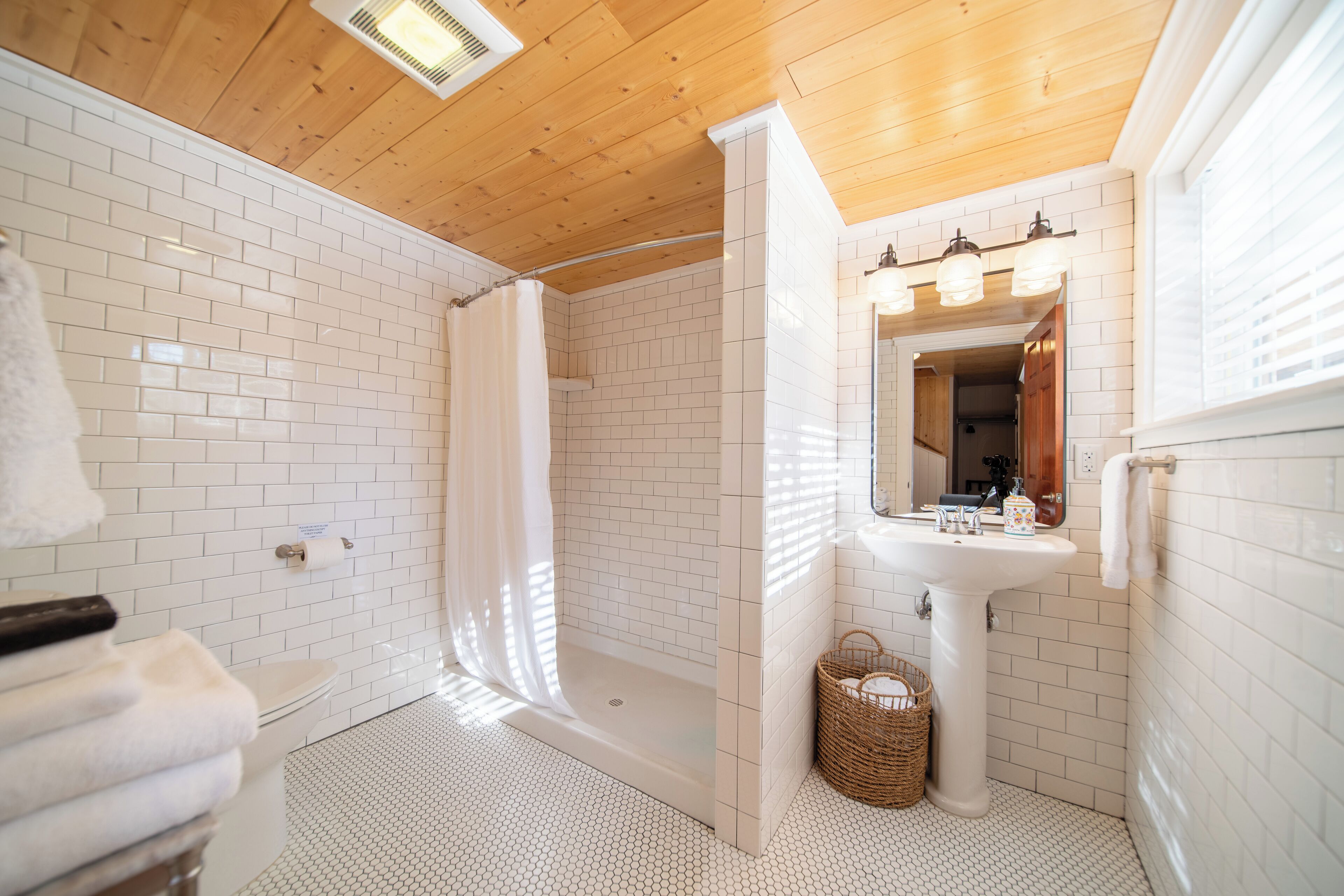 A bathroom with white tiled walls, a wooden ceiling, a white sink, a mirror, and a shower area.