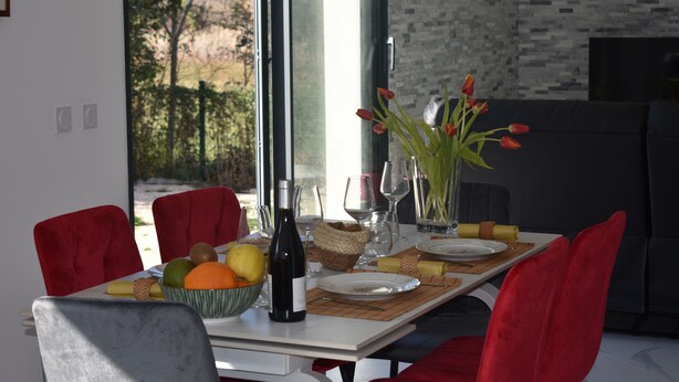 Une table à manger dressée avec un saladier de fruits, une bouteille de vin et des verres, entourée de chaises rouges et offrant une vue sur les arbres à l’extérieur.