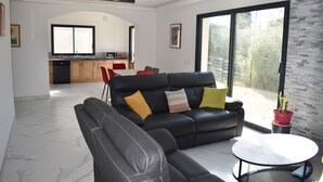 Un salon moderne avec un canapé en cuir noir, une table basse ronde et un coin repas comprenant une chaise rouge et une cuisine en arrière-plan.