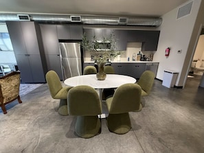 A modern kitchen with a stainless steel refrigerator, a dining area with a round table and green chairs, and a potted plant on the table.