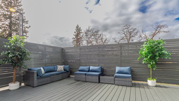 A covered outdoor seating area with a dark-colored wooden floor, a grey woven sofa set with blue cushions, and a potted plant.