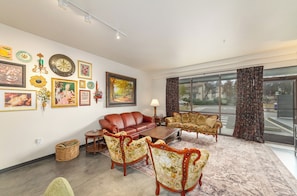 A spacious living room with a leather sofa, floral-patterned armchairs, a wooden coffee table, and a large window with curtains.