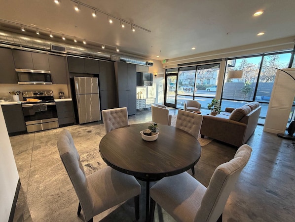 A modern kitchen with stainless steel appliances, a dining area with a round table and cushioned chairs, and a seating area with sofas and a large window offering a view of the street.