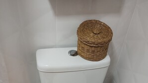 Eine Toilette mit einem gewebten Korb auf dem Deckel, einer kleinen Topfpflanze daneben und einem Holzregal darüber.
