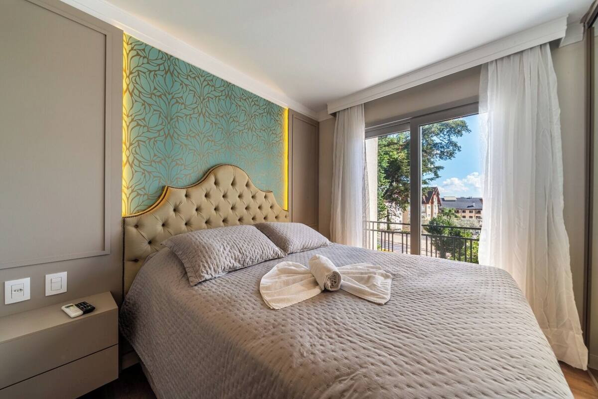 A hotel room with a large bed, a headboard with a floral pattern, a bedside table with a remote control, and a view of the outside through a balcony.