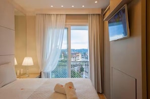 A hotel room with a bed, a nightstand, a lamp, a window with curtains, and a view of buildings. - Stay Home Luxo Gramado | 1min Rua Coberta | Ar Cond | Premium (Gramado)