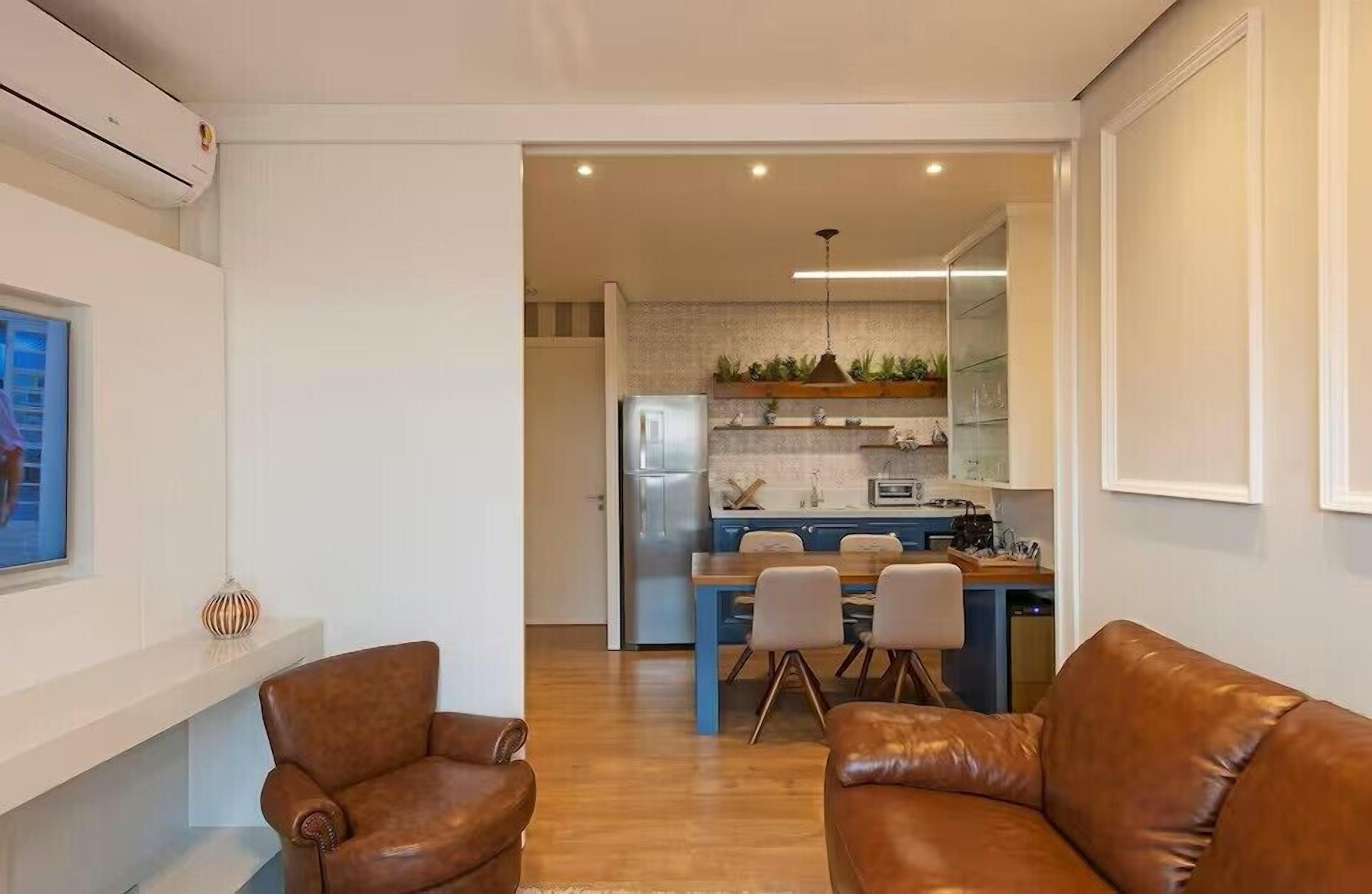A modern living room with leather sofas, a dining area with a blue table, and a kitchen with a built-in fridge and plants.