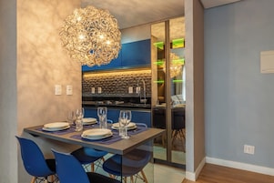 Uma área de jantar moderna com uma mesa posta para quatro pessoas, um candelabro e uma cozinha ao fundo.