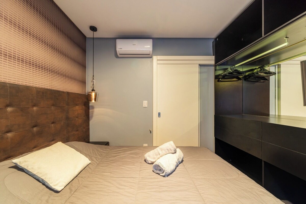 Camera d'albergo moderna con un letto, condizionatore d'aria a parete, lampadario e uno specchio che riflette un portascarpe.