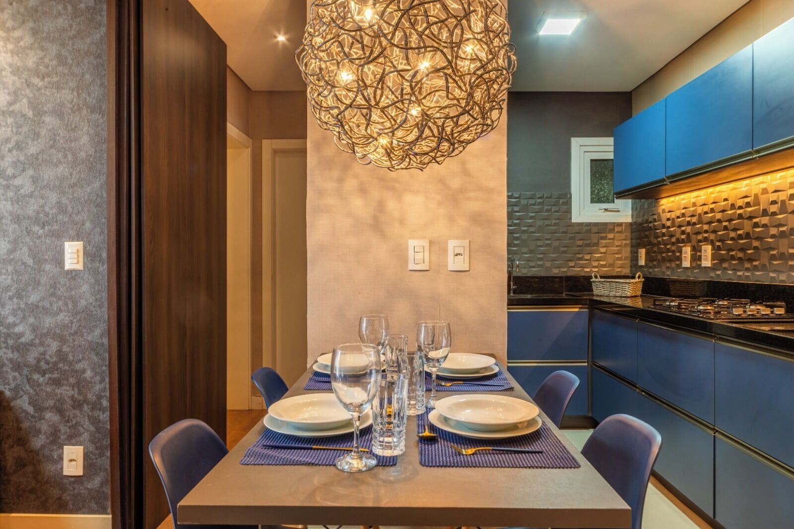 A modern dining area with a table set for four, a hanging light fixture, and a kitchen with blue cabinets.