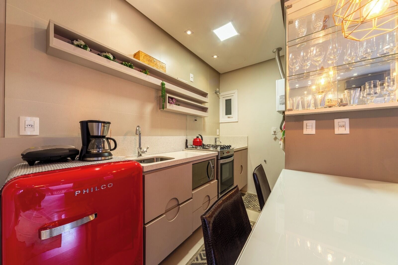 Una cocina compacta con un refrigerador PHILCO rojo, una cafetera, una estufa a gas y un lavamanos. Hay estantes sobre la encimera y una vitrina de vidrio con vasos.
