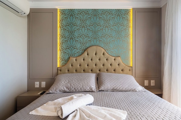 A hotel room with a tufted headboard, a bed with two pillows, a folded towel, and a patterned wall panel. - Stay Home Luxo Gramado | 1min Rua Coberta | Ar Cond | Premium (Gramado)