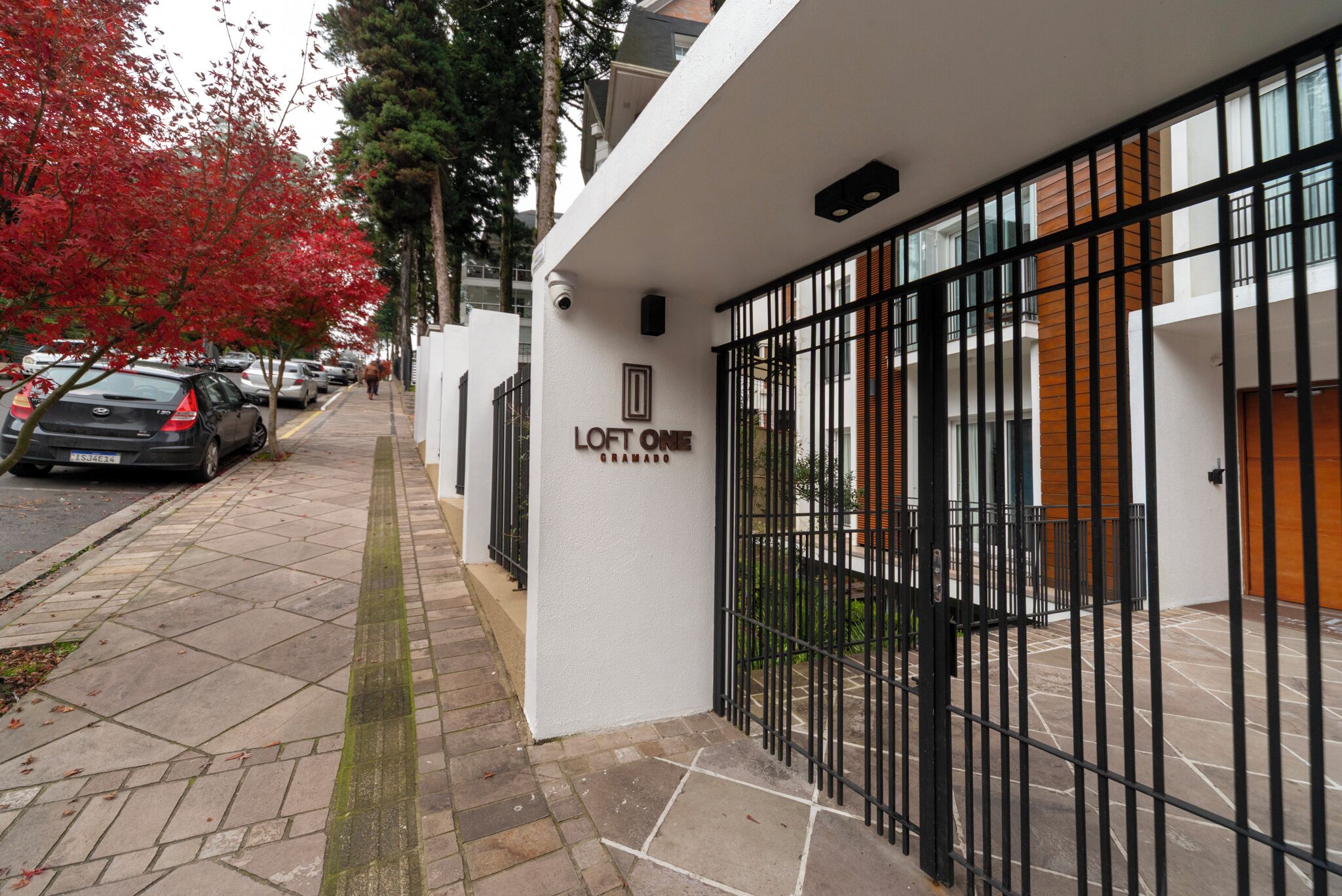 Un bâtiment moderne doté d’une entrée sécurisée par un portail, d’une enseigne indiquant « LOFT ONE », et d’un trottoir pavé où sont garées des voitures.