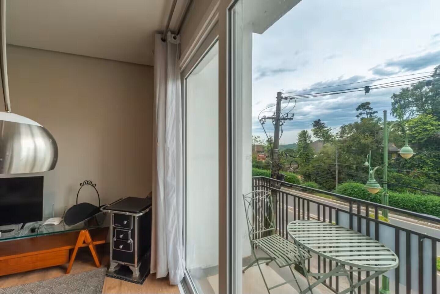 A balcony with a metal table and chairs, a TV on a stand, and a view of greenery and power lines.
