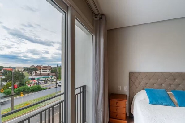 A hotel room with a large window, a bed with blue pillows, and a wooden nightstand.