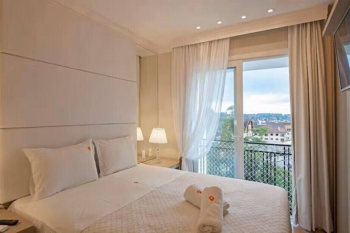 A hotel room with a bed, a nightstand, a lamp, a window with curtains, and a view of buildings.