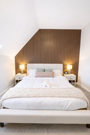 A modern bedroom with a sloped ceiling, a large bed with white bedding, two bedside tables with lamps, and a wooden panel wall. - King Spacious Yale Getaway near Wooster Sq Park (New Haven)