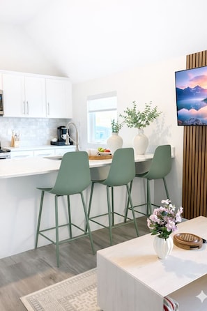 A modern kitchen with white cabinets, a bar area with green chairs, a coffee machine, and a large window. - King Spacious Yale Getaway near Wooster Sq Park (New Haven)
