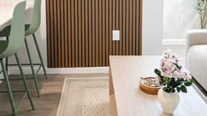 Ein modernes Wohnzimmer mit einem Flachbildfernseher an einer Wand aus Holzpaneelen, einem Essbereich mit grünen Stühlen, einem Couchtisch mit Blumenvase und Blick auf Berge durch das Fenster.