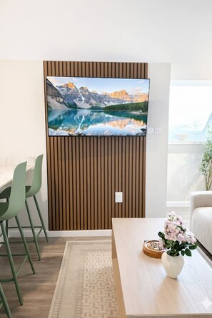 A modern living room with a flat-screen TV mounted on a wooden panel wall, a dining area with green chairs, a coffee table with a vase of flowers, and a view through a large window. - King Spacious Yale Getaway near Wooster Sq Park (New Haven)