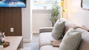 Ein modernes Wohnzimmer mit einem großen Flachbildfernseher an der Wand, einer Couch mit Kissen, einem kleinen Tisch mit Blumen in einer Vase und einem Fenster mit Blick ins Freie.