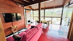 Een moderne woonkamer met een rode bank, houten wanden, een flatscreen televisie en grote ramen met uitzicht op de natuur.