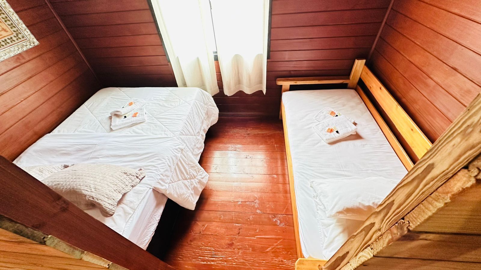 Una habitación pequeña y acogedora con dos camas de madera, cada una con una colcha blanca y un cojín pequeño. La habitación tiene paredes y suelo de madera.