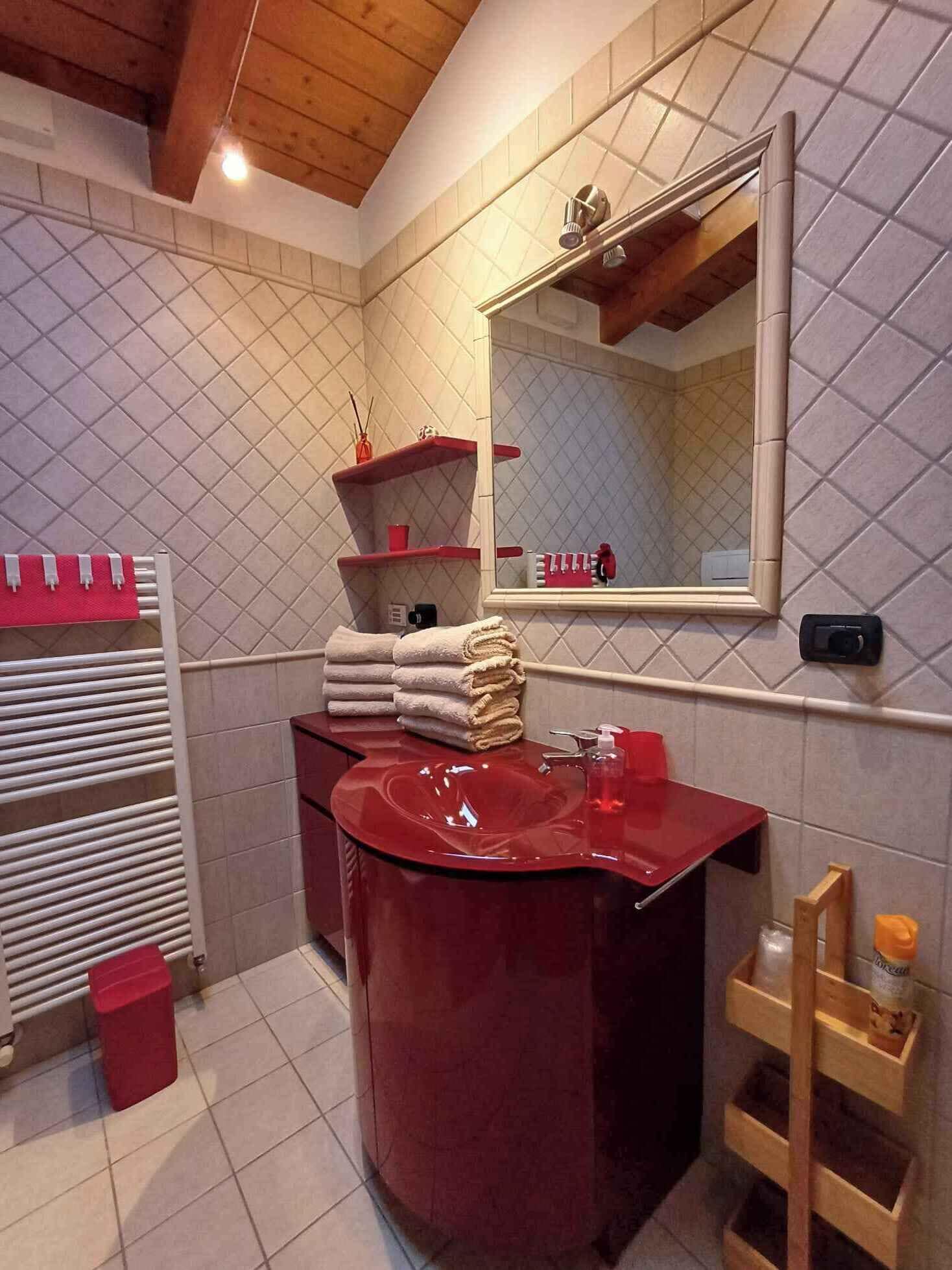 Un bagno con un lavandino rosso, uno specchio, un porta asciugamani e uno scaffale in legno.