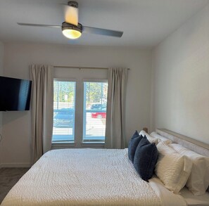 Un dormitorio con cama, ventilador de techo, televisor montado en la pared y ventana con cortinas.