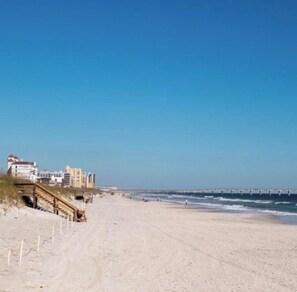 A beach with a wooden boardwalk leading to buildings, a clear sky, and a calm sea.