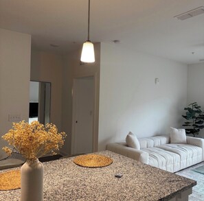 A modern living room with a white sofa, a dining area with a granite table, and a hanging pendant light. - Azure Luxe Haven Near TPC, Mayo Clinic & Beaches! (Jacksonville)