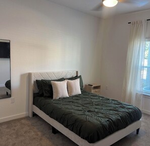 A bedroom with a bed, a nightstand, a mirror, and a window with curtains. - Azure Luxe Haven Near TPC, Mayo Clinic & Beaches! (Jacksonville)