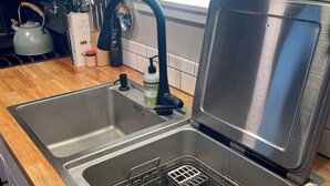 En stålvask med en opvaskestativ, en elkedel og en flaske opvaskesæbe på bordpladen.