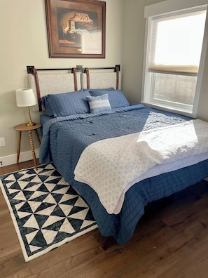 A bedroom with a bed, a nightstand, a lamp, a framed picture on the wall, and a window.