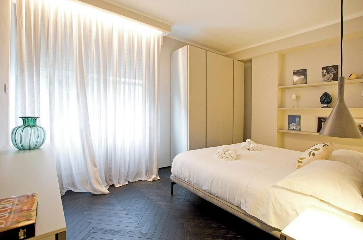 A hotel room with a bed, a wardrobe, a desk with a vase, and a window with curtains.