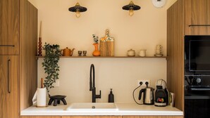 Une cuisine avec des meubles en bois, un plan de travail blanc, un évier, une bouilloire, une machine à café et une plante suspendue.