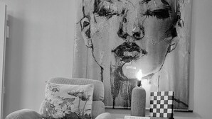 Une chambre moderne avec un grand tableau abstrait au mur, un fauteuil confortable agrémenté d’un coussin à motifs floraux, une petite table sur laquelle repose une boîte à carreaux, et un luminaire suspendu.