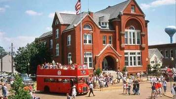 A red double-decker bus in front of a large brick building with arched windows and a flagpole.