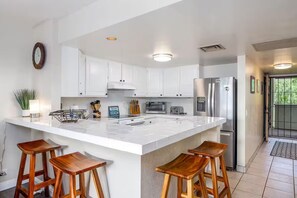 A modern kitchen with white cabinets, a large island with stools, stainless steel appliances, and a clock on the wall. - Central AC Ocean View 2bedroom /2bath Sleeps 6 Condo across Magic Sands Beach (Kailua-Kona)