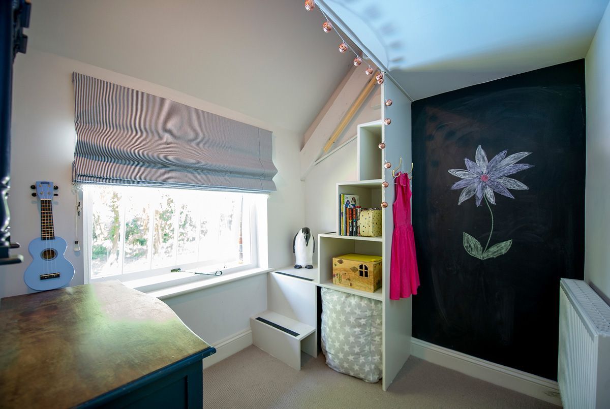 Une chambre d’enfant avec une fenêtre, une guitare, une bibliothèque et un mur orné d’un tableau représentant des fleurs.