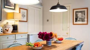 Ein Holztisch mit einer Tomatenschale und einer Blumenvase. Über dem Tisch hängen zwei Lampen, und im Hintergrund ist eine weiße Tür zu sehen.