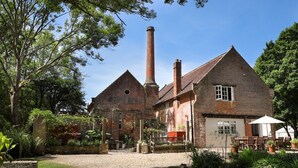 Ein traditionelles Backsteingebäude mit einem großen Schornstein, einer überdachten Terrasse mit Sitzgelegenheiten und einem Garten.