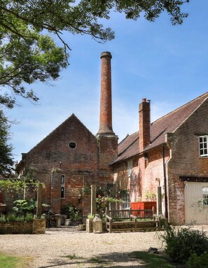 A brick building with a tall chimney, a circular window, and a wooden bench in the courtyard. - The Brick House & Annexe (Beaulieu)